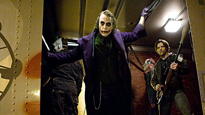 Heath Ledger as the Joker in The Dark Knight pointing a gun with city chaos in the background.